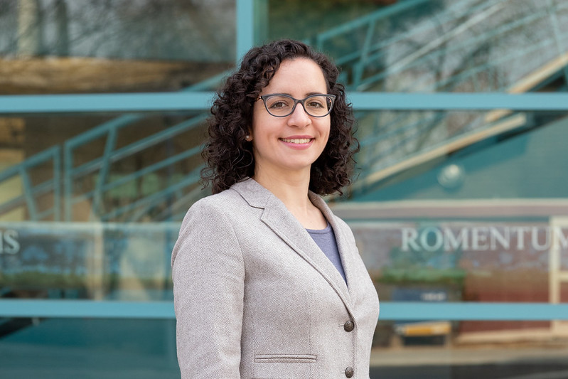 Young white woman with curly hair and glasses and a sweater standing in front of a UCD building