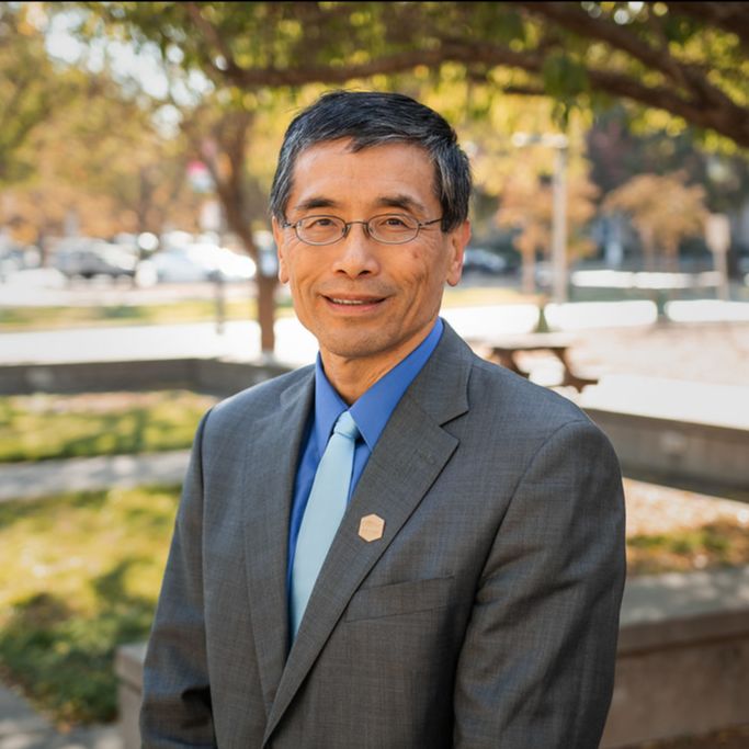 Middle aged Asian man in a gray suit with a blue tie standing outside under a tree