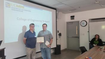 People in a classroom, one accepts an award and a projected slide on the wall has the award name
