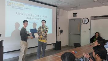 People in a classroom, one accepts an award and a projected slide on the wall has the award name