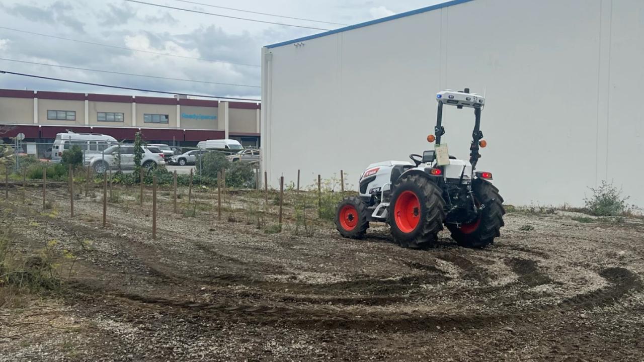 An Agtonomy-enabled Bobcat CT4045 autonomous tractor operates on a dirt lot beside a large building, with stakes marking rows and parked vehicles visible in the background under a cloudy sky.