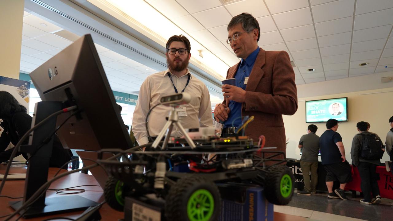 Two men stand in front of a computer with a robotic vehicle in the foreground