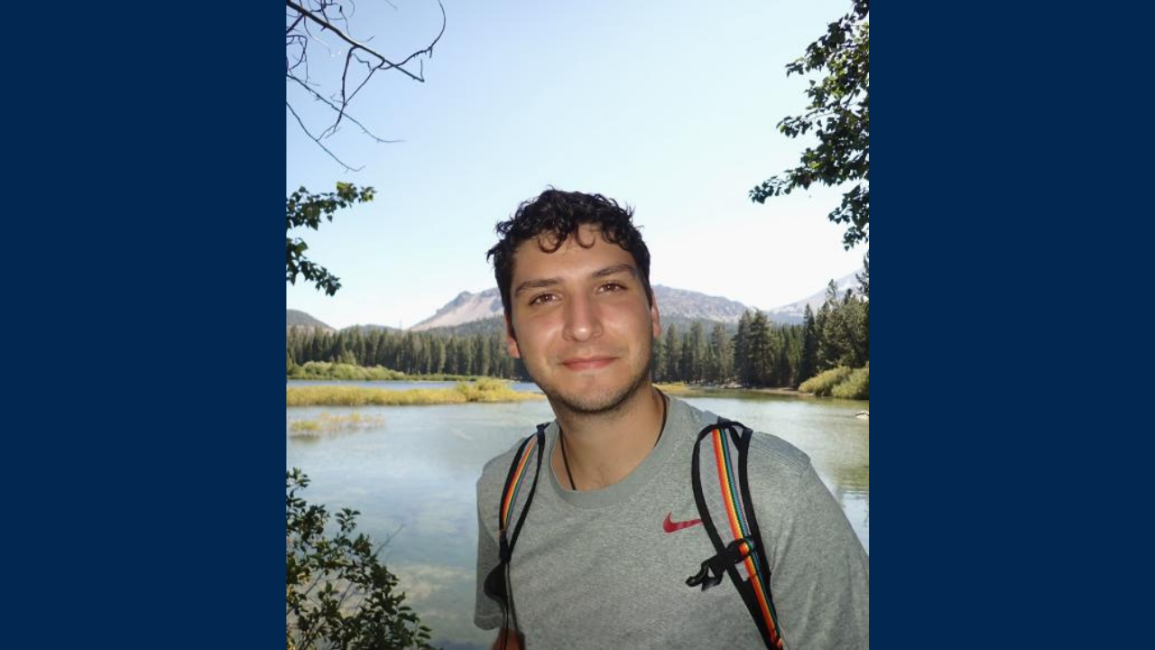 Gonzalez with backpack smiling by a mountain lake and pine trees