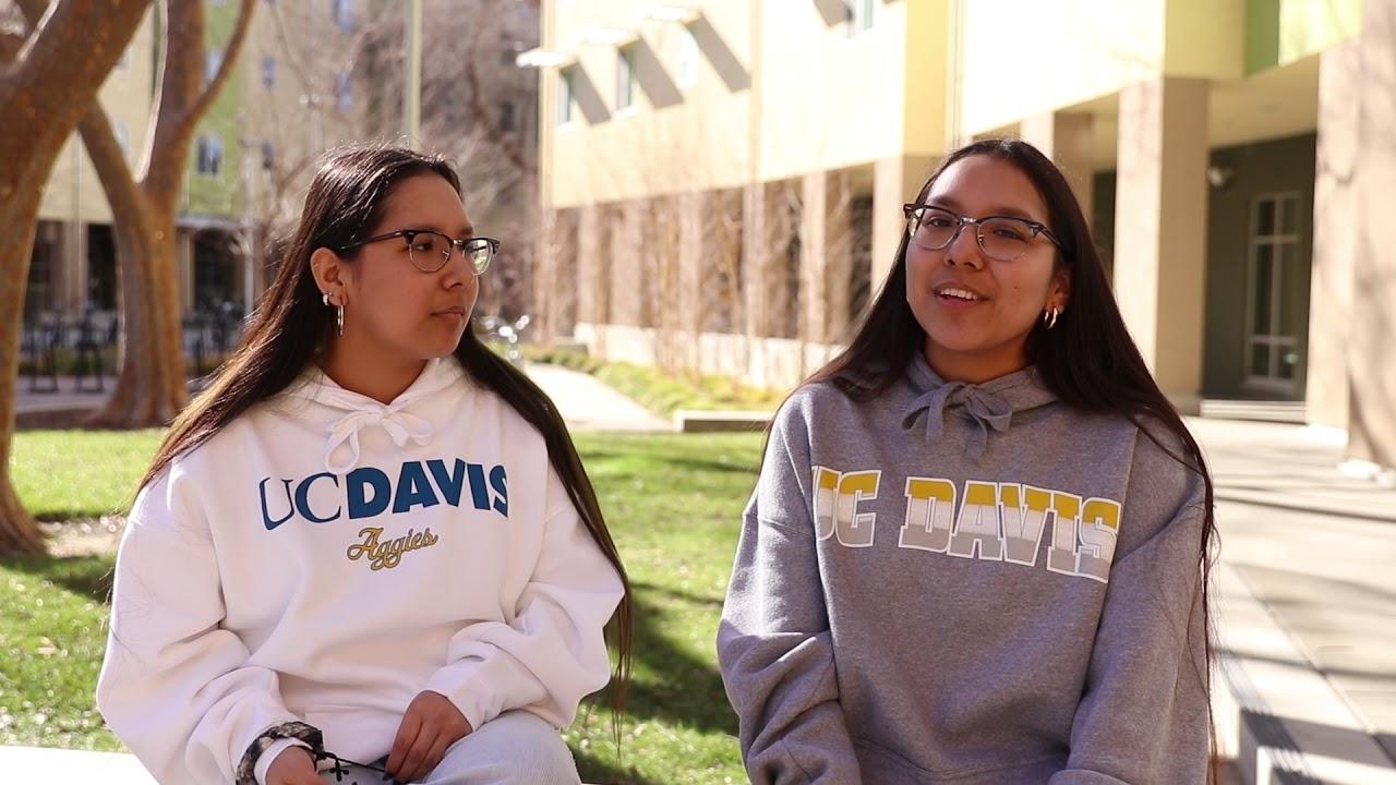 Twins in UC Davis sweaters sit outside near a building