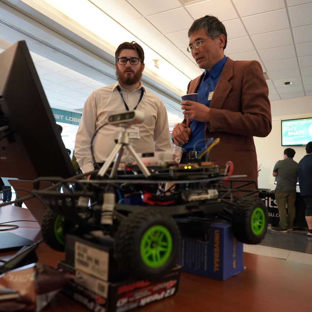 Two men stand in front of a computer with a robotic vehicle in the foreground
