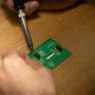 A green microchip sitting on a wooden table is worked on by a pair of hands holding a tool