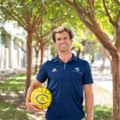 Smiling student in navy polo holding yellow water polo ball on sunlit tree-lined walkway