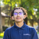 Headshot of young man with glasses, curly hair, navy polo, slight smile, blurred green background