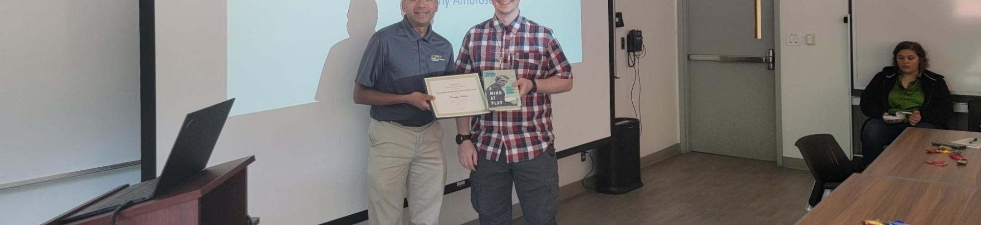 People in a classroom, one accepts an award and a projected slide on the wall has the award name